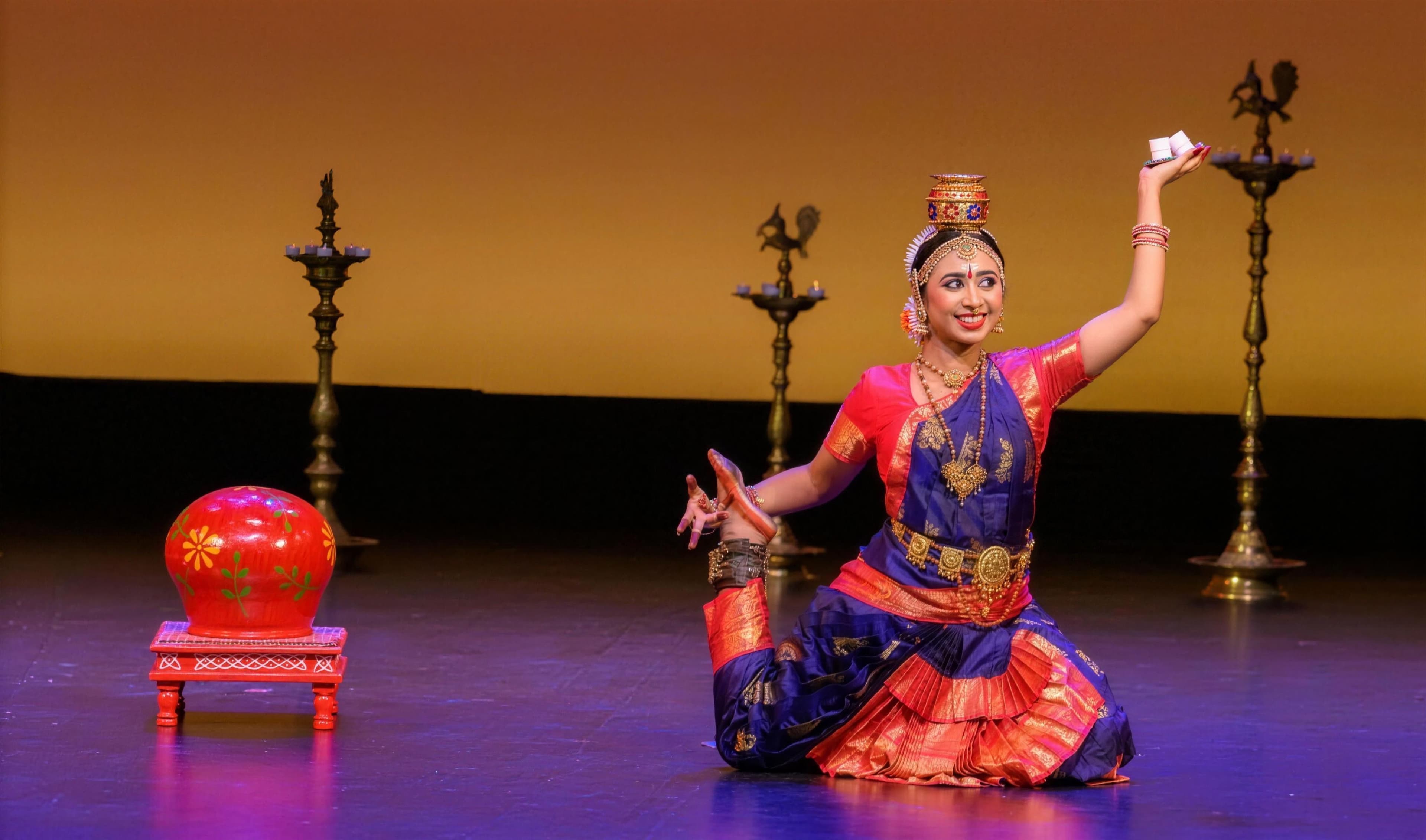 A student kneeling onstage in a red and blue Bharatanatyam costume, a kalasam balanced on her head, one hand raised in a mudra beside a row of lit brass diyas.