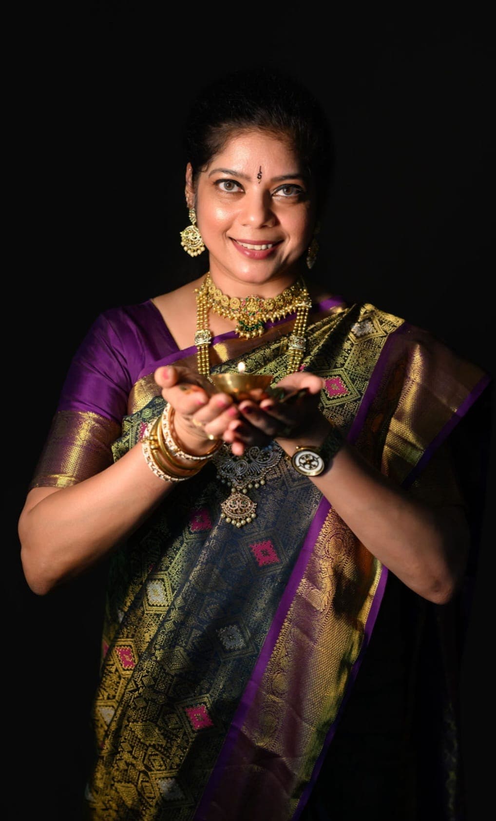 Chandna Talluri in a purple and gold silk saree, holding a lit brass diya in both hands.